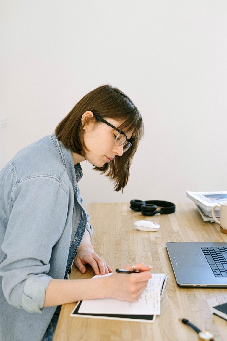 Femme concentrée qui prend des notes dans un bureau cosy avec ordinateur portable, écriture de blog